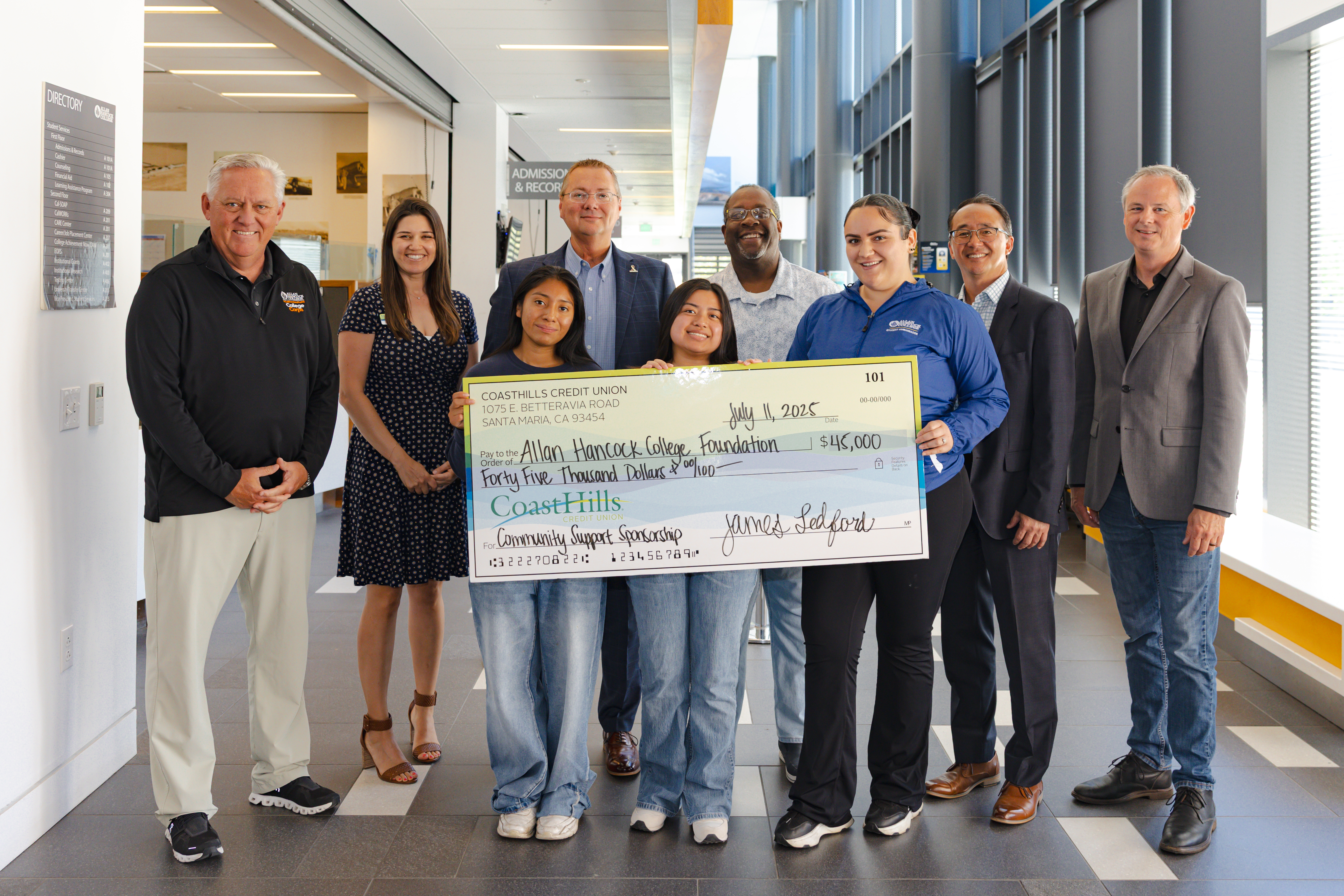 Representatives from Allan Hancock College, CoastHills Credit Union, and Hancock students pose together holding an oversized ceremonial check for $45,000 from CoastHills Credit Union to the Allan Hancock College Foundation. The group stands in a bright campus hallway near the Admissions & Records office